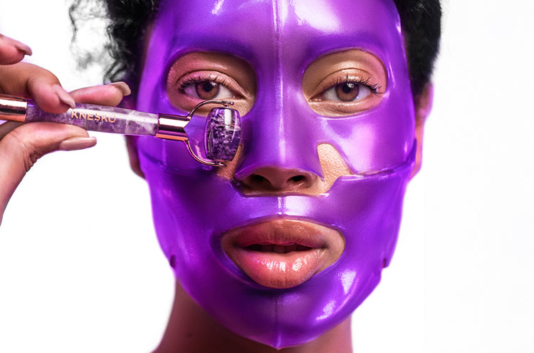Woman wearing a purple face mask and using a jade roller on her face.